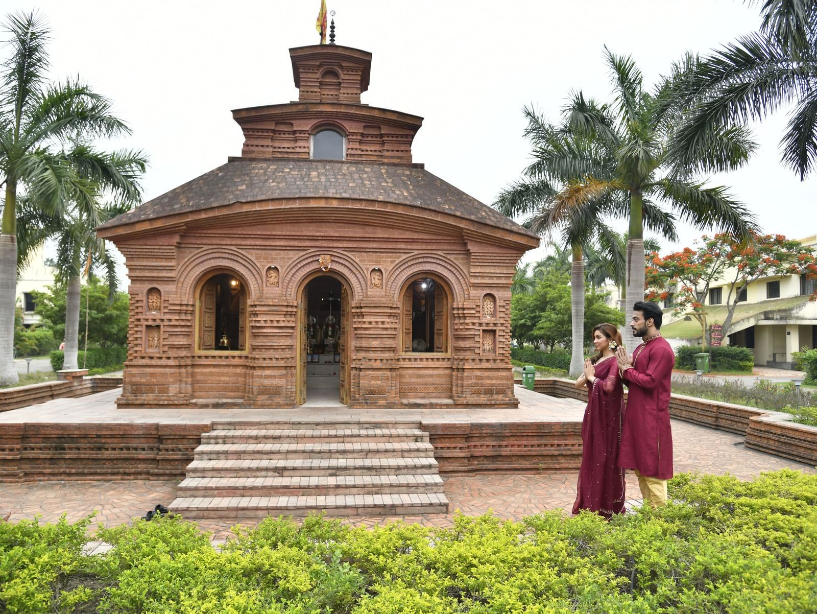 A multi-tiered brick temple-like structure shows stairs leading up to it, with a couple in traditional attire standing at the base and palm trees surrounding it