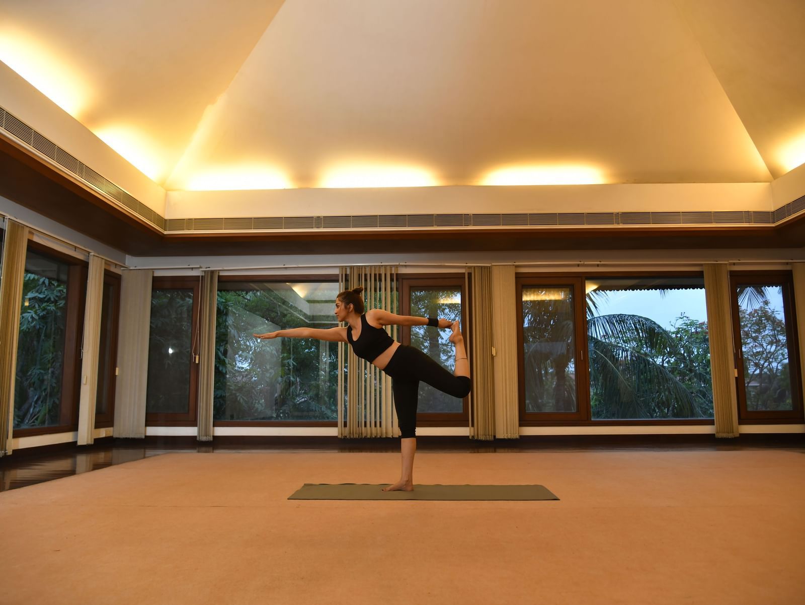 A person performs a yoga pose on a mat in a spacious, well-lit room with large windows and warm-toned lighting