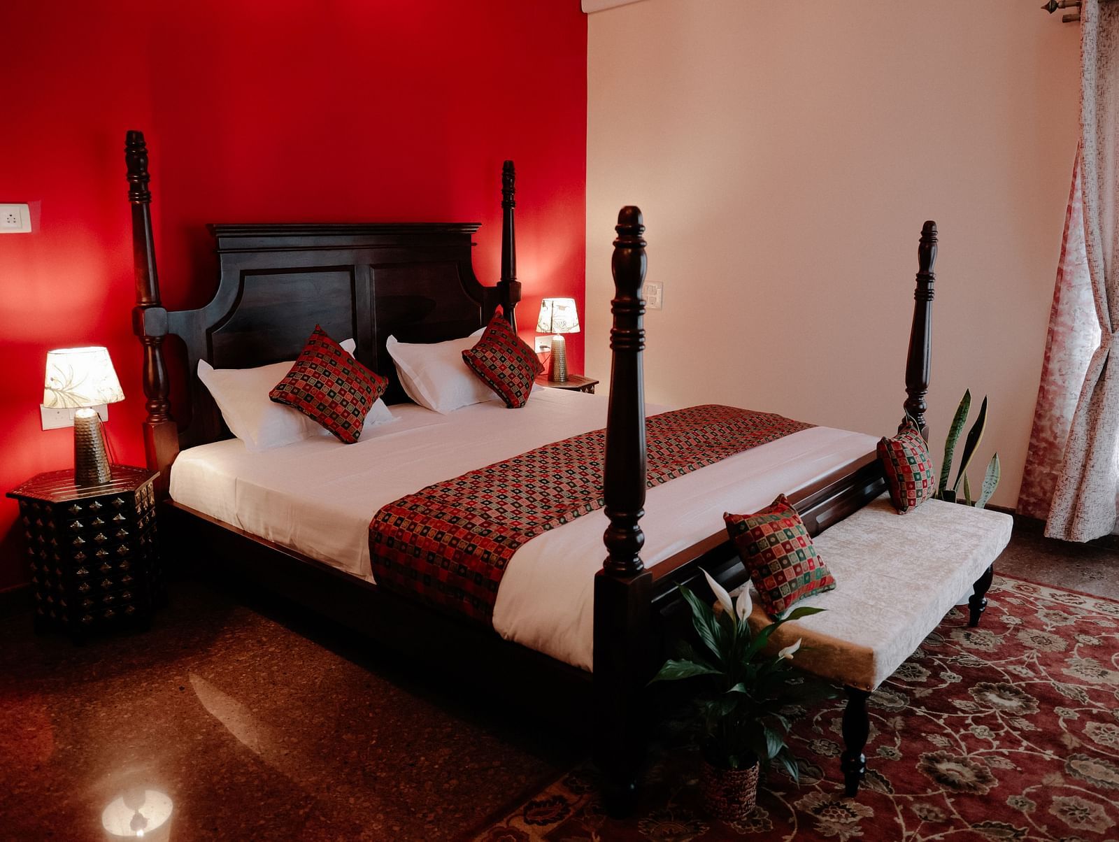 A dark wood four-poster bed with white linens and red patterned throw pillows in a room with a bold red accent wall and a lighter adjacent wall | Adhyaya Heritage Reverbed