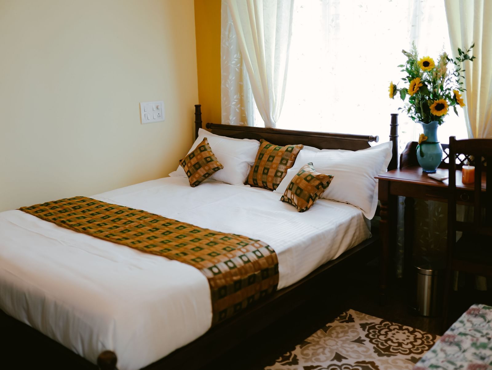 A bedroom with a wooden bed with white linens and patterned throw pillows and a throw blanket, next to a small table | Adhyaya Heritage Reverbed