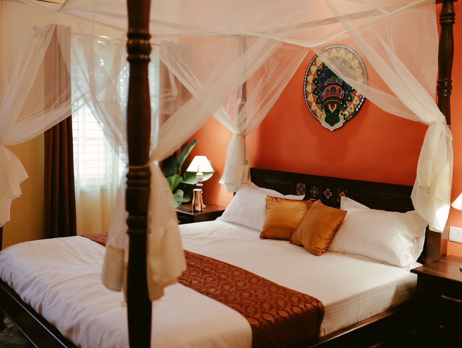 A four-poster bed with white drapery and a decorative headboard in a room with an orange accent wall | Adhyaya Heritage Reverbed
