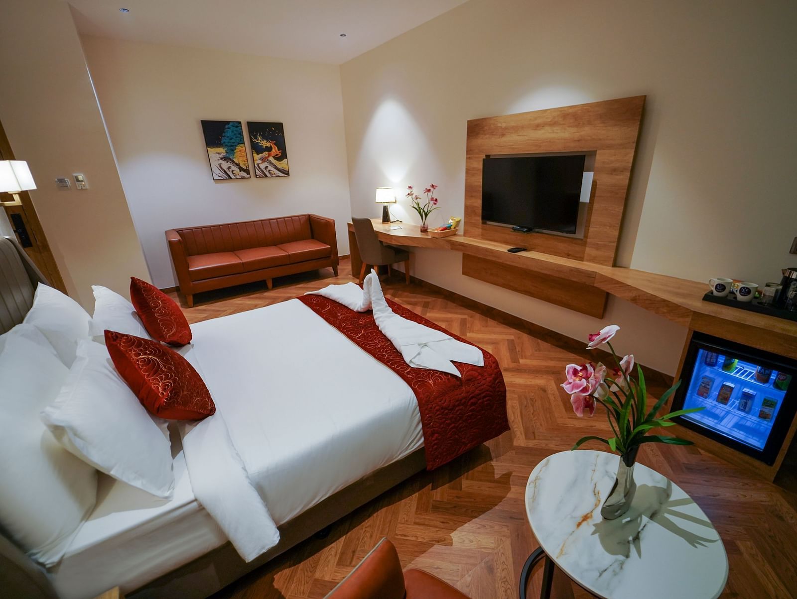 A hotel room offers a bed with pillows, a sofa, a desk area with a lamp and flowers, and two televisions - Hotel Park Inn, Nellore.