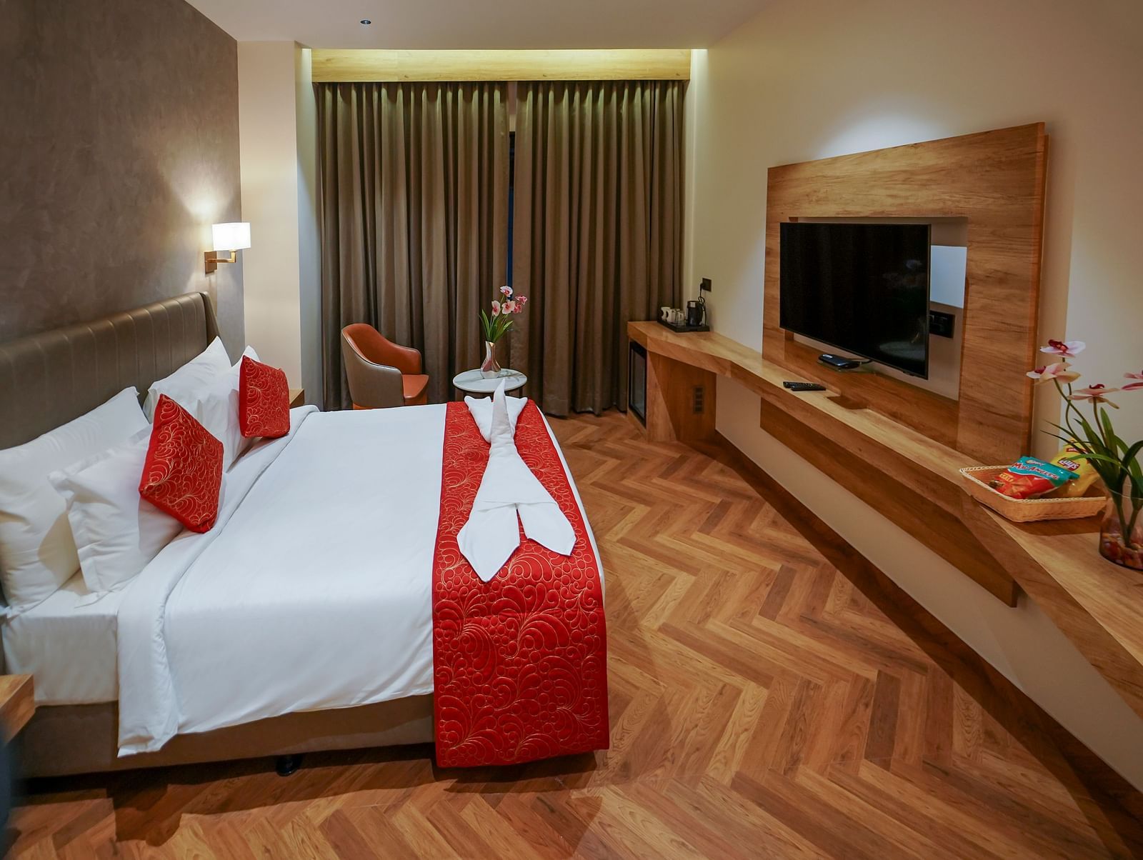 A hotel room features a bed with pillows and folded linens, curtains, a mounted television, and a long desk area with flowers - Hotel Park Inn, Nellore.