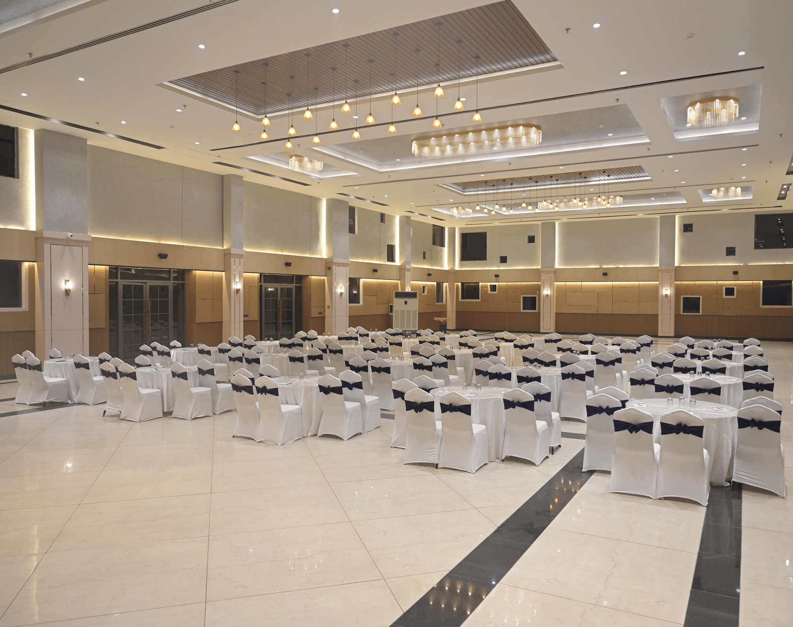 A panoramic view of the banquet hall arranged for a gathering, featuring bright chandeliers and neatly placed round tables at Hotel Sonar Bangla Mayapur.