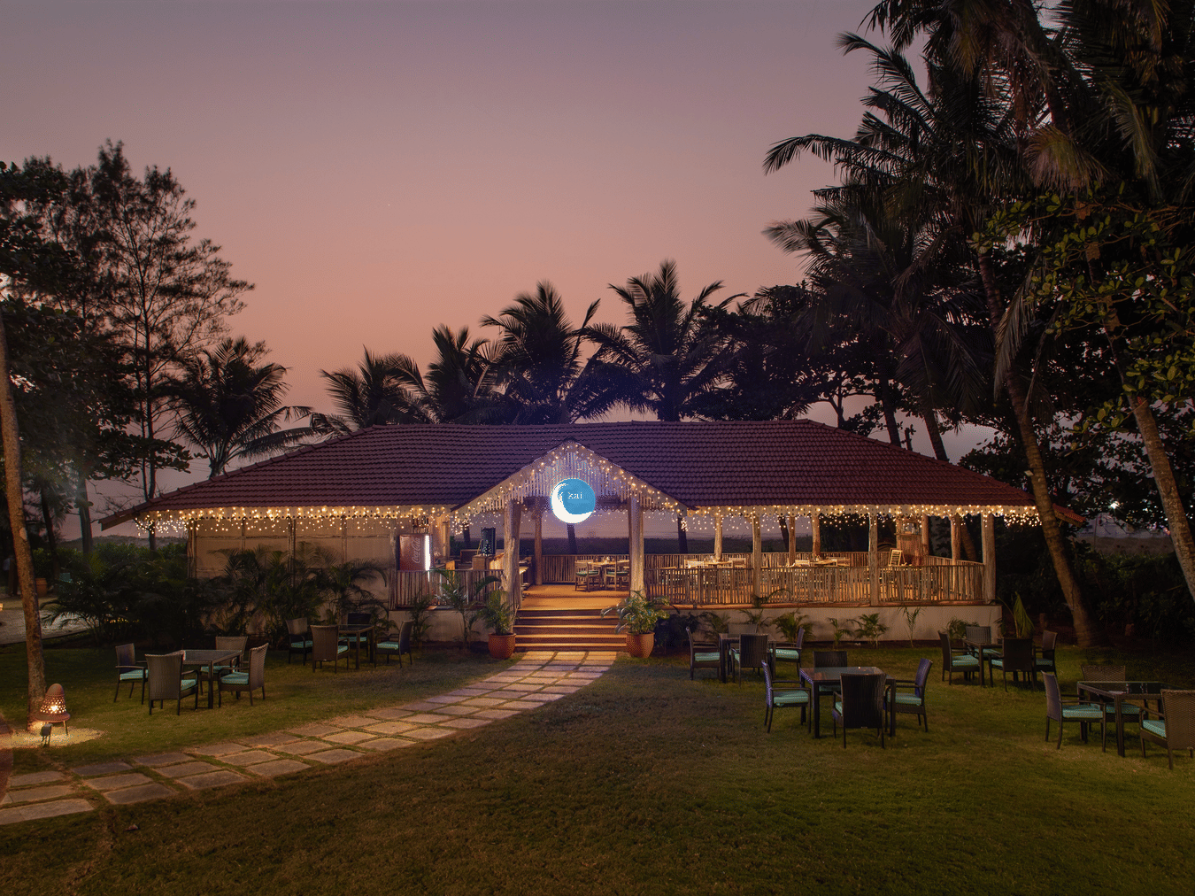 The exterior facade of Kai, the seaside restaurant that looks enchanting in the late evening sky with a walkway leading to the restaurant and trees surrounding the exterior at Kenilworth Resort & Spa, Goa.