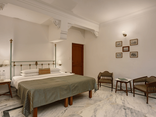 A  bedroom at Ram Pratap Palace, Udaipur, featuring a well made four-poster bed, two wooden chairs, a small table, and framed art on the wall.