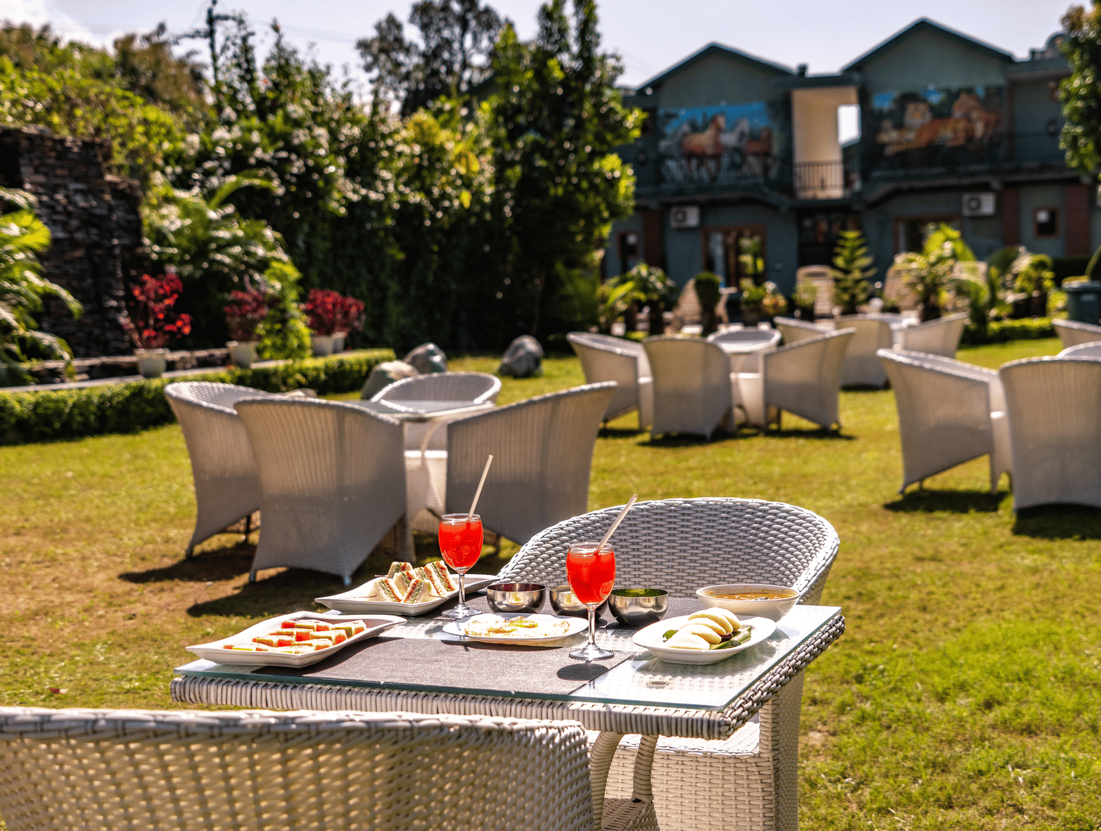 Outdoor dining setup at Corbett Nirvana Resort on a lawn, featuring white woven tables and chairs amidst the manicured garden.