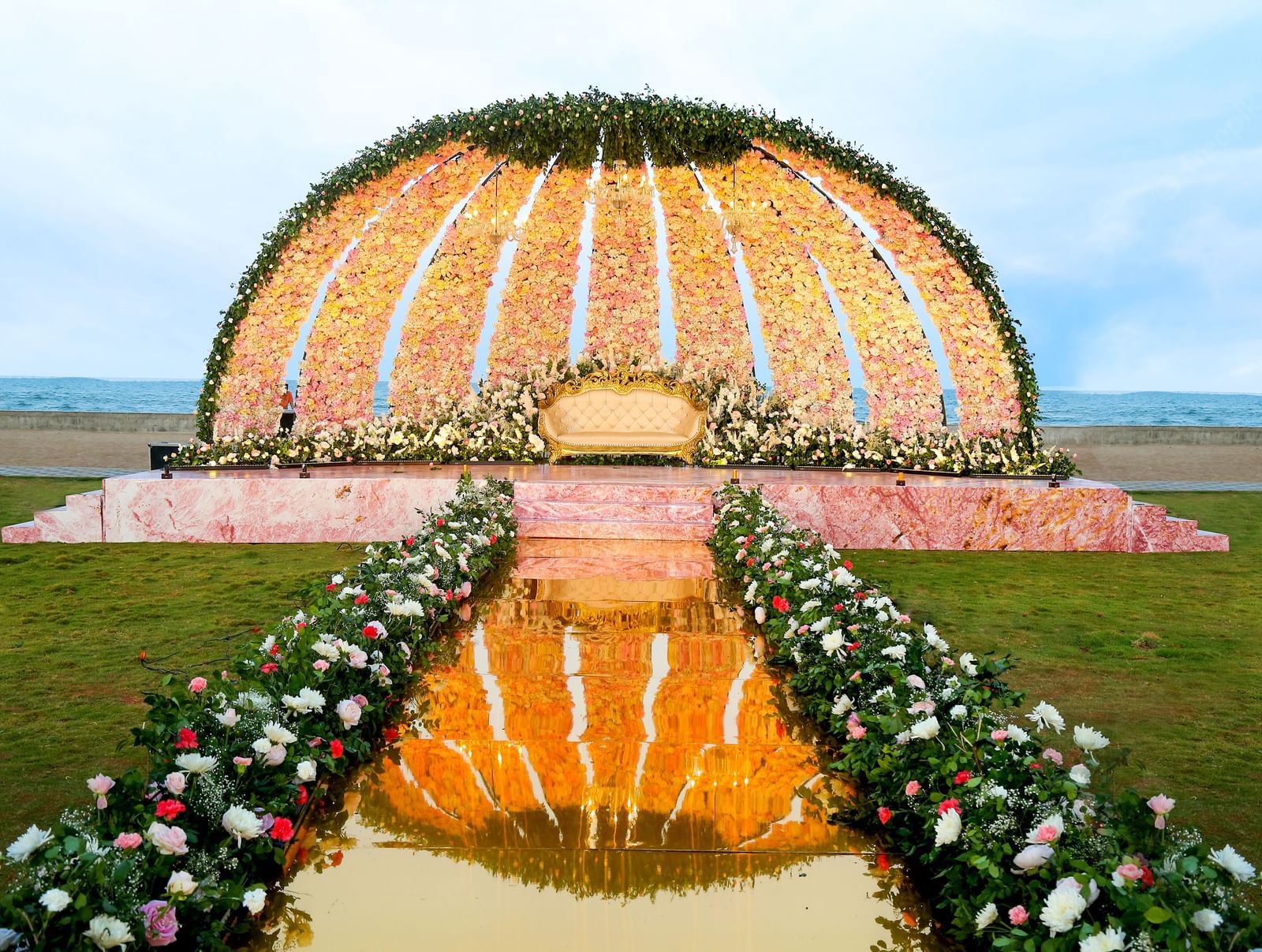 A large, decorative outdoor archway made of white and orange flowers and fabric, leading to a path on a beach at sunset | Horizon | Grand Bay Resort and Spa