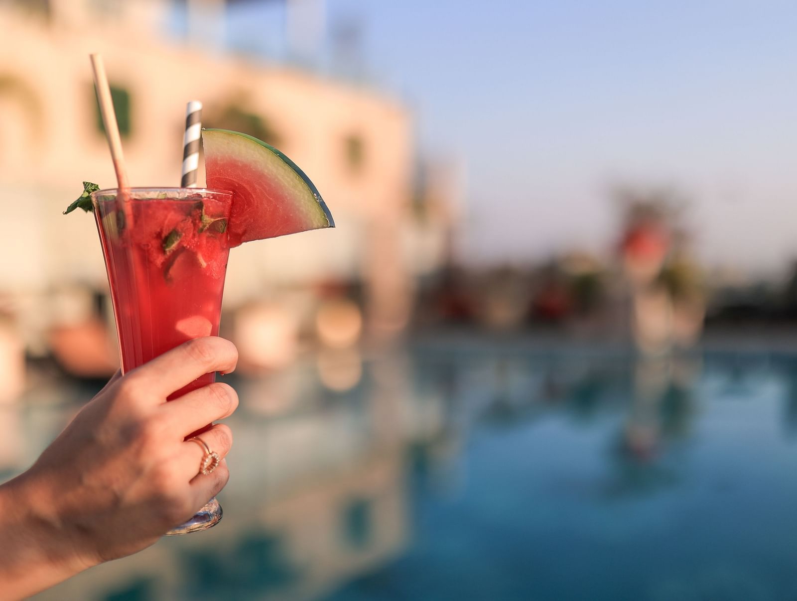 A person's hand holding a tall glass of watermelon juice, garnished with a slice of watermelon and a straw, against a blurred background.