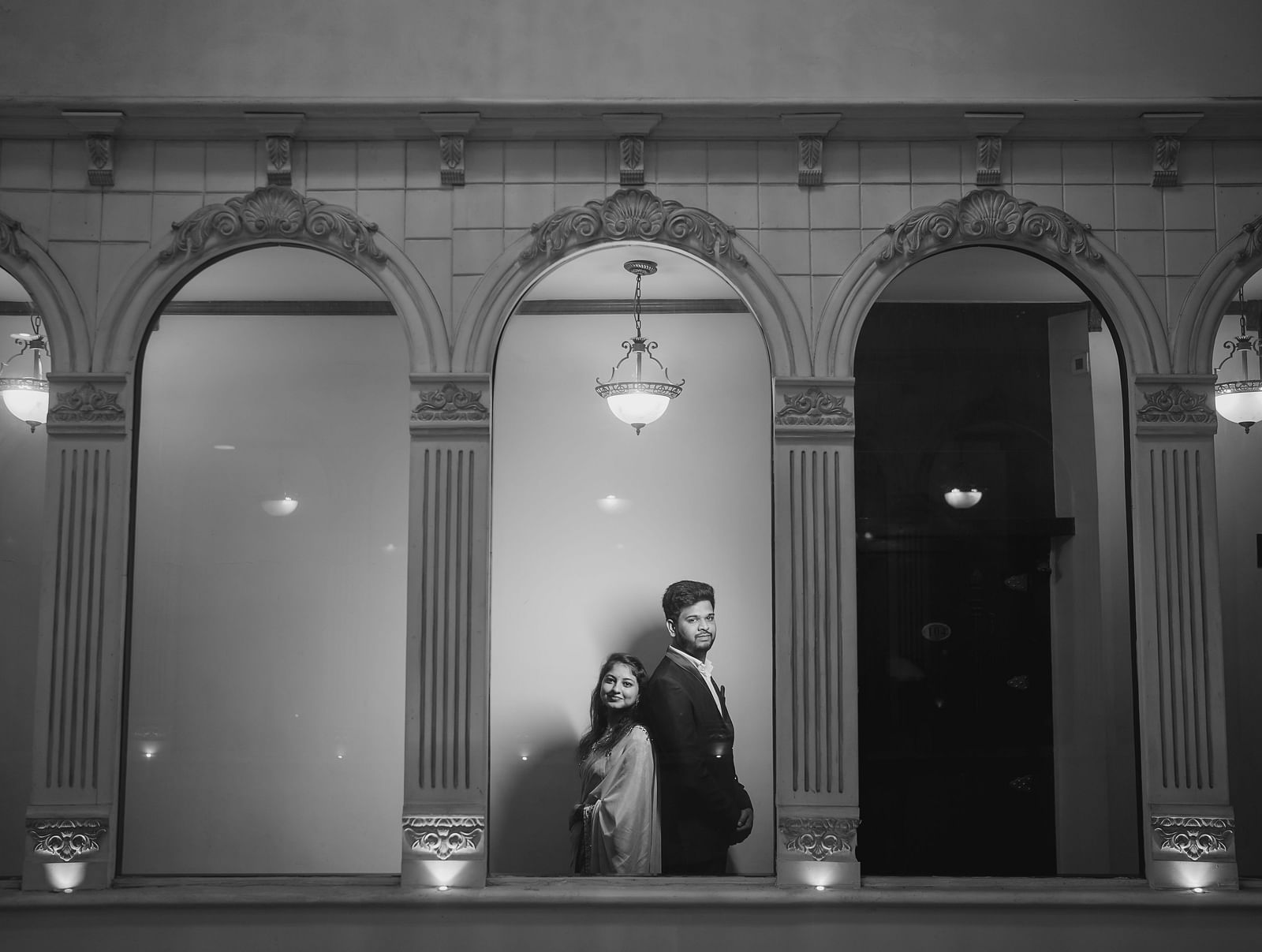 A monochrome shot features a couple embracing within the arched openings of a grand, classic building.