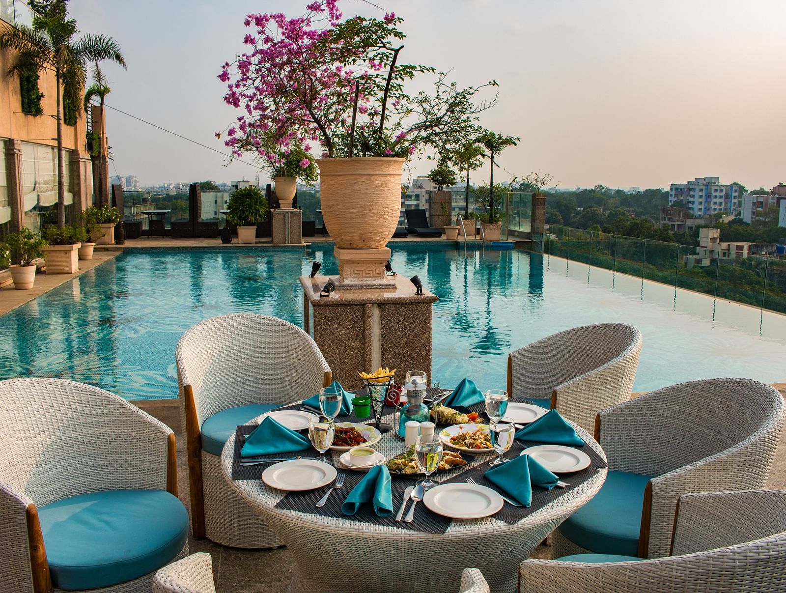 An outdoor dining at Hotel Ashok Nagpur featuring a round table with place settings for four, surrounded by cushioned chairs, and a swimming pool and cityscape in the background. 