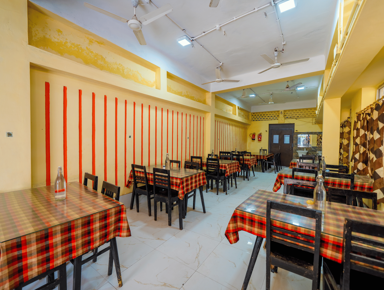 A brightly-lit, empty hotel dining hall with small tables covered in red and black checkered tablecloths and matching chairs - Hotel Dolphin Puri
