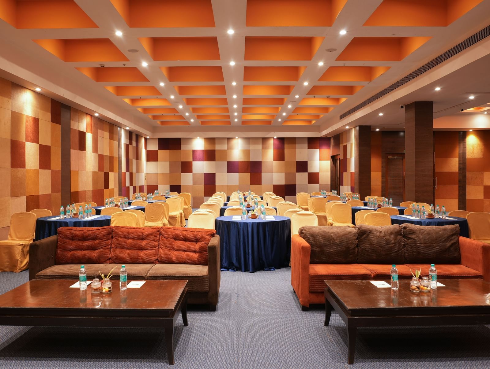 Modern interiors of V-Den at Vedic Village Spa Resort featuring brown plush couches, neatly arranged banquet tables, and elaborative ceilings