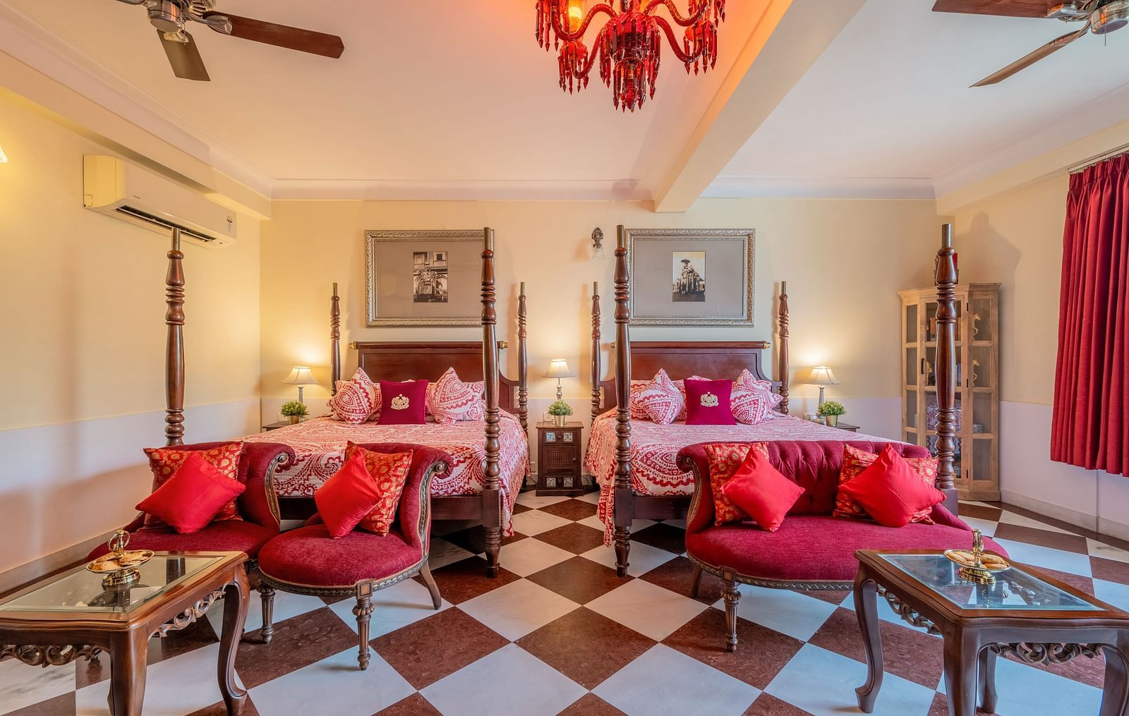A vibrant suite at Dera Rawatsar, Jaipur, features two elegant four-poster beds, comfortable seating areas, ornate tables, and striking checkered flooring.