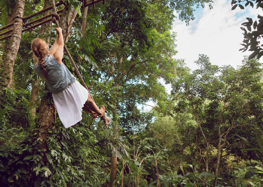 A girl in a frock swinging a tarzan swing, tied to a tree top, in a dense forest.