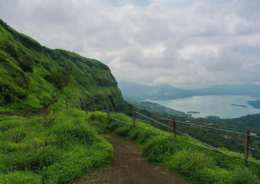 Lush green hillside with a narrow path curving along the slope, overlooking a distant lake and cloudy sky, conveying a sense of tranquil exploration.