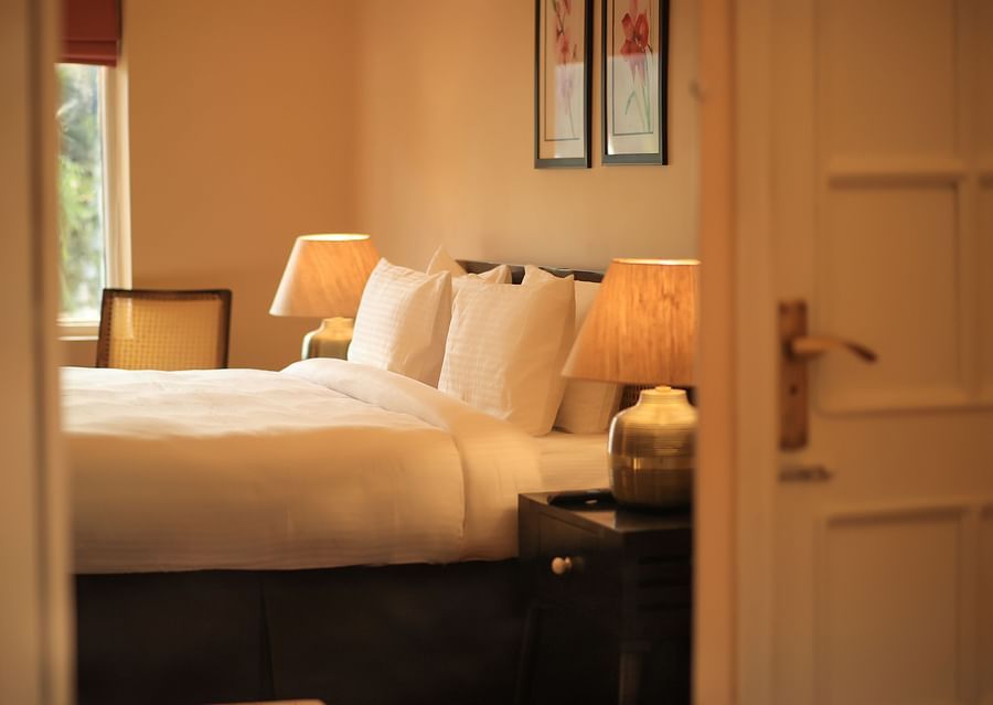 A glimpse of the Super Deluxe Room 4 where a bed with pillows, an open window, and a bedside table with lamp can be seen - Lamrin Boutique Cottages Rishikesh