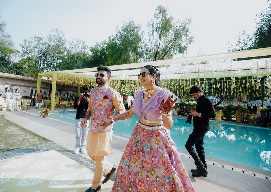 Bride and groom in coordinated floral outfits walking beside a pool at a daytime event.