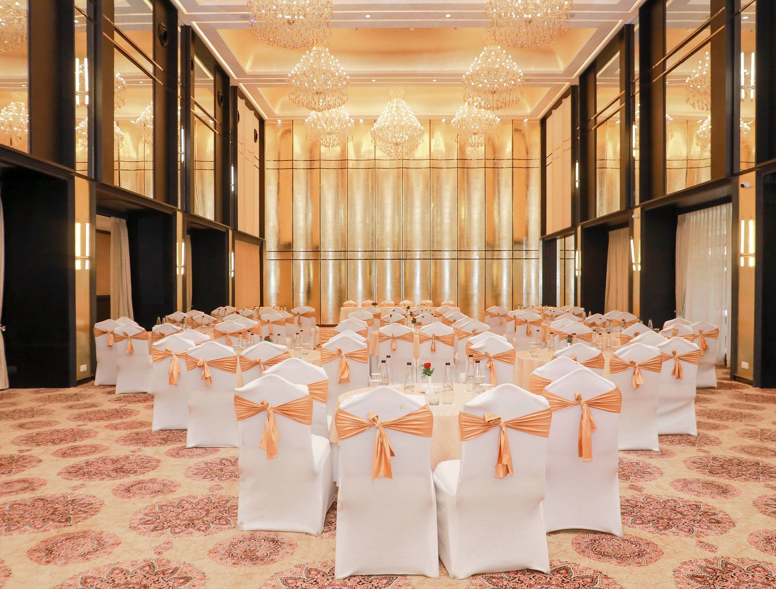 Banquet hall set up for an event, featuring white chairs with peach sashes, grand chandeliers, high ceilings, and warm lighting.