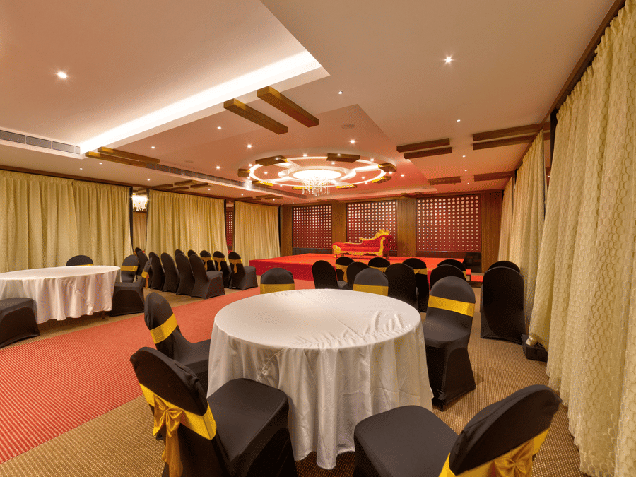 Round tables, black and gold chairs, a red carpet, and a stage setup inside the banquet hall of Icon Express By Bhagini, Mahadevapura.