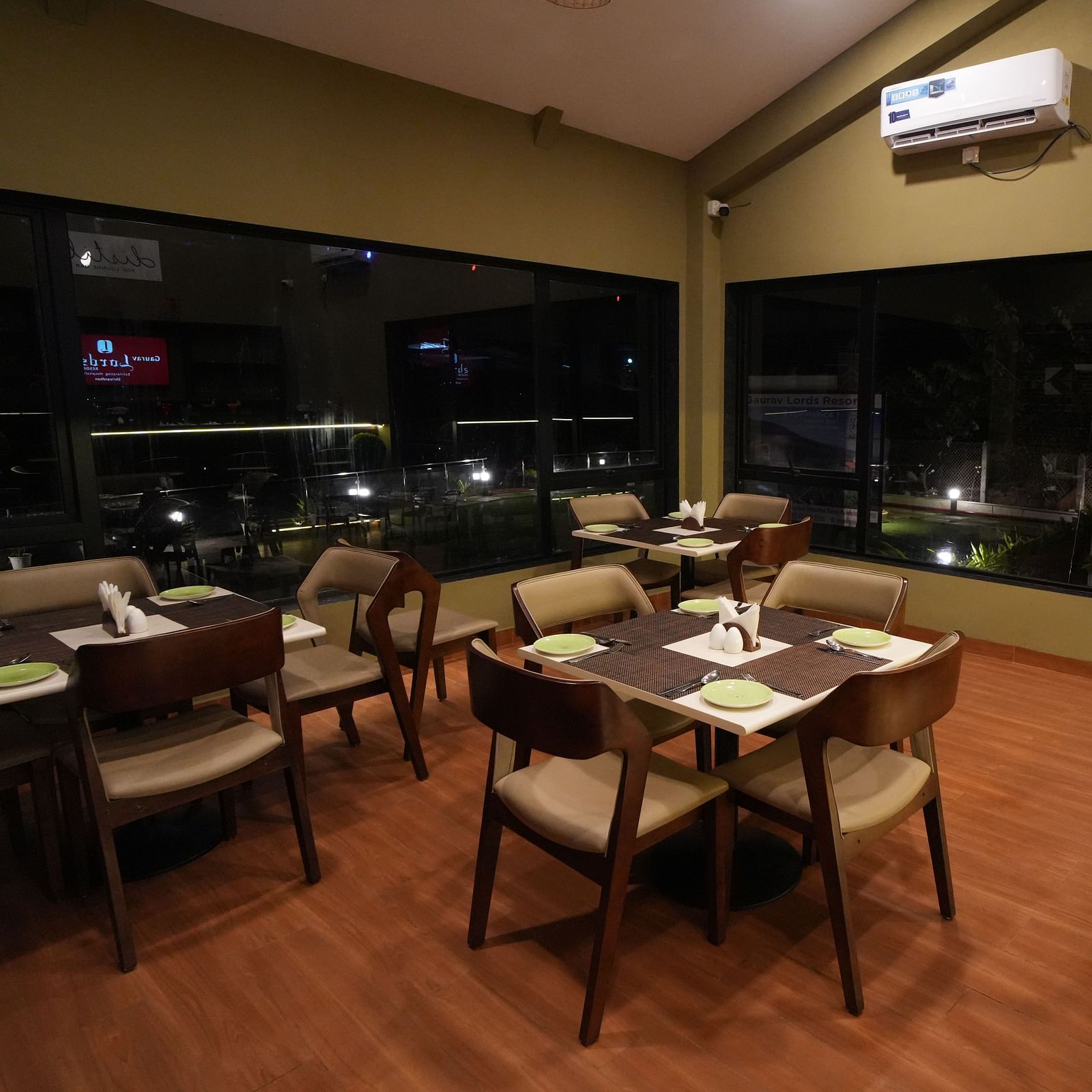 Cosy indoor dining area with modern wooden tables and cushioned chairs, large glass windows offering night views, and bar stools lined along a dark counter - Gaurav Lords Resort Shrivardhan