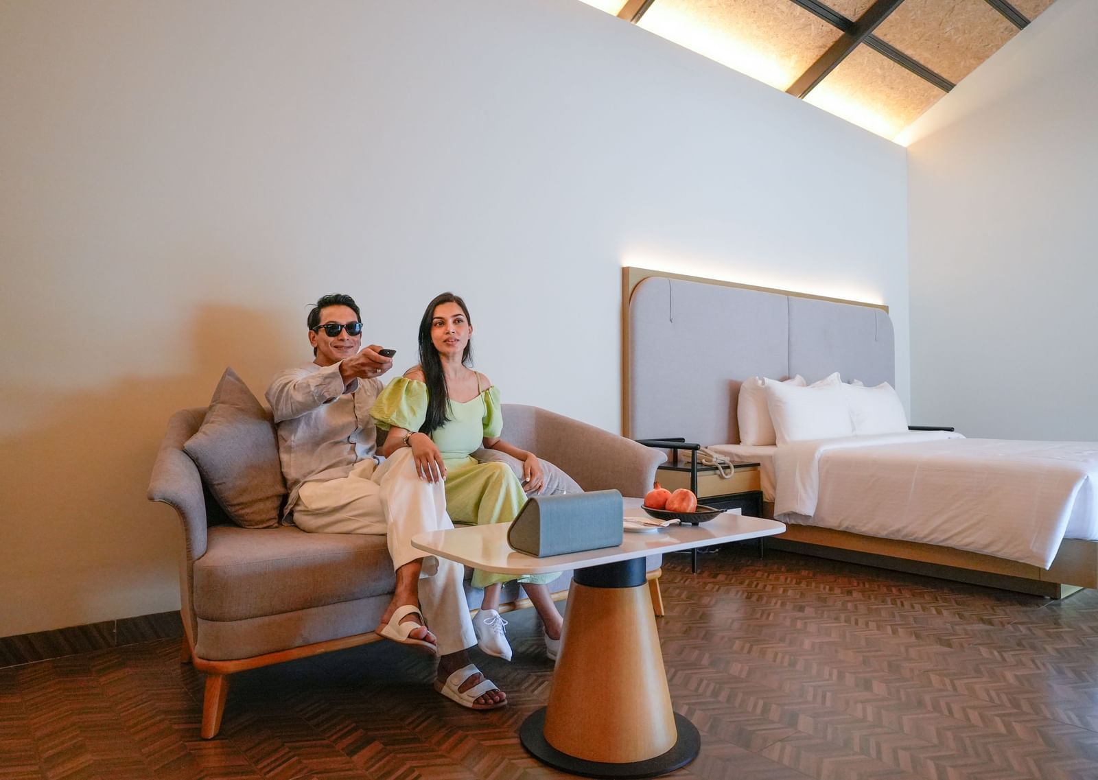 Couple relaxing on a sofa in a modern hotel room with a bed, coffee table, and warm lighting - The Shore Trishvam, Pondicherry