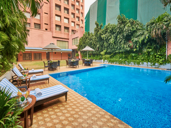 An outdoor swimming pool during the day with blue water, lounge chairs, and a multi-story building in the background | HHI Varanasi