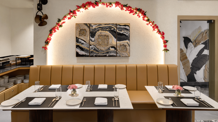 An indoor restaurant with a long, padded banquette seat against the wall, individual tables with place settings, and a decorative archway with red flowers and a piece of abstract wall art | Voyage Eastin Suites