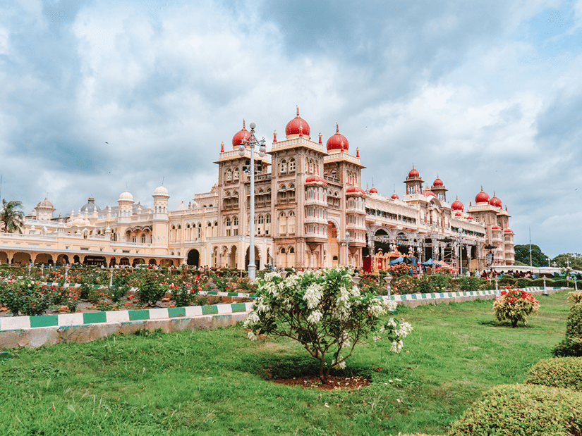 The exterior of the Mysore Palace with its ornate walls and architecture with greenery in the foreground under a clear blue sky.