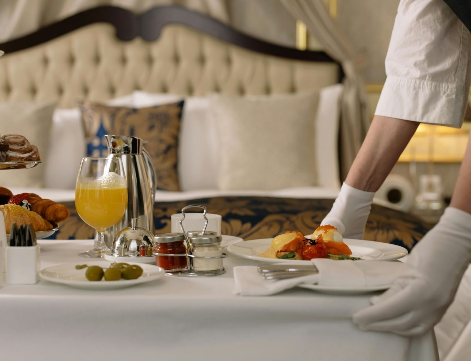 A pair of hands setting up a breakfast tray with orange juice and food on a table in front of the bed.