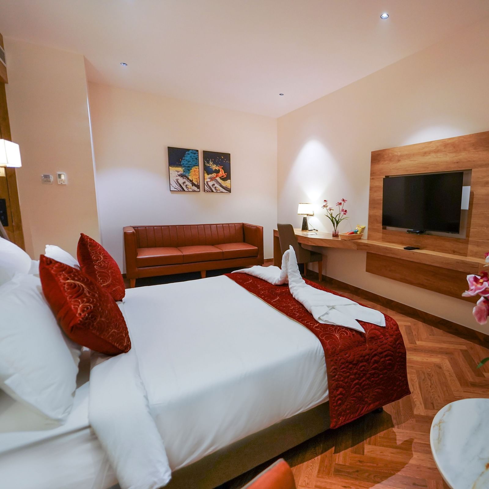 A hotel room includes a bed with pillows, a sofa, a desk with a lamp and flowers, and two televisions - Hotel Park Inn, Nellore.