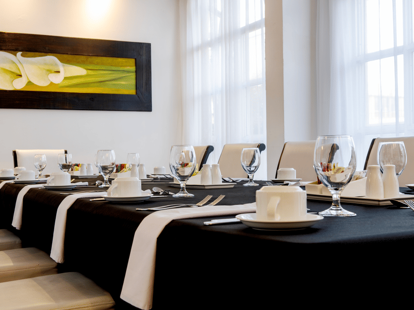 A dining table set with plates, folded napkins, glasses, and cutlery beside large windows inside the Boardroom of S Hotel Kingston.