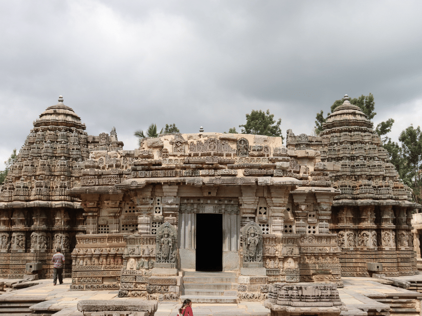 Historic stone temple featuring intricately carved pillars, ornate towers, and traditional South Indian architecture beneath a cloudy sky.