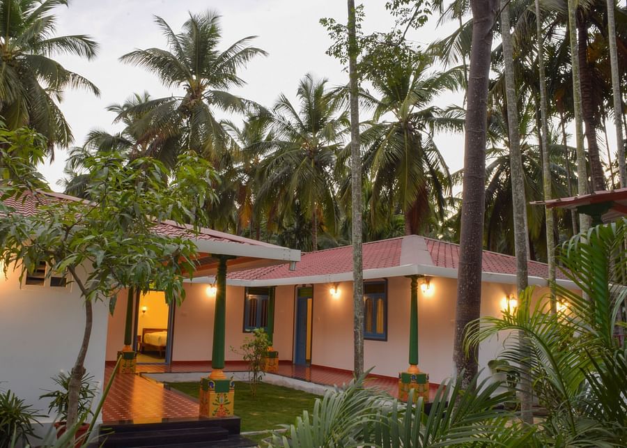 A white resort cottage with a red roof and blue window frames, surrounded by palm trees and green lawns - Ibex Resorts, Coimbatore (Kakarla)