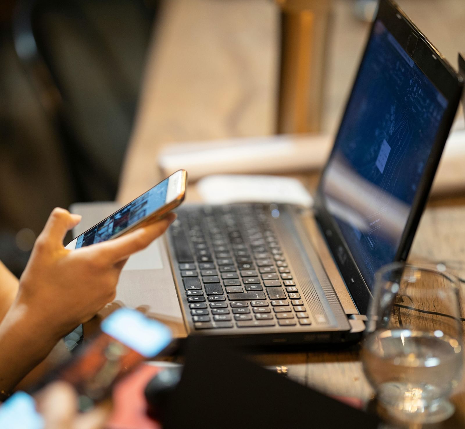 A laptop kept on a table beside a half-empty glass in front of a woman scrolling through her phone.