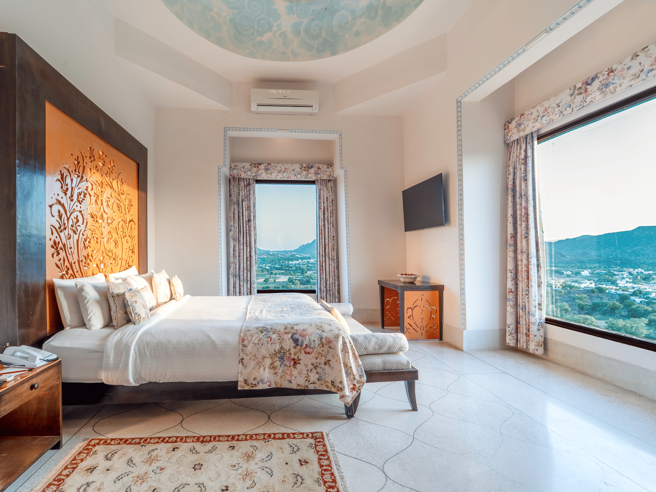 A grand bedroom with a king size bed, ornate headboard, and a large window overlooking the Aravalli hills - Fateh Garh, Udaipur