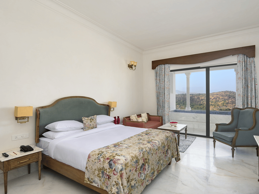 Interior view of the Plunge Pool Suite featuring a queen size bed, with side tables, lamps, a chair and a floor to ceiling sliding door to reach the plunge pool, offering the finest accommodation in Udaipur