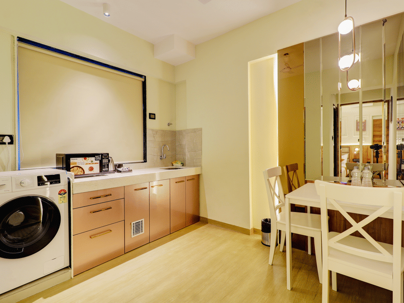 The interior at  Home2 India, with washing machine, cabinets, countertop, and sink, with a small dining table and chairs placed near a glass partition.