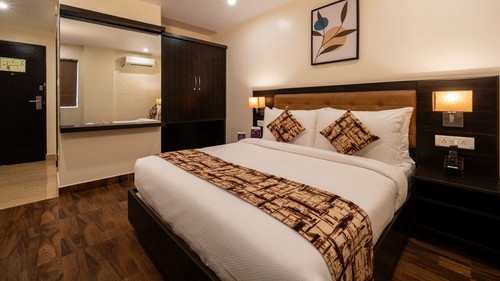 A bedroom interior at VITS Select, Bengaluru, featuring a plush bed, wooden flooring, and a bathroom entrance with mirror.