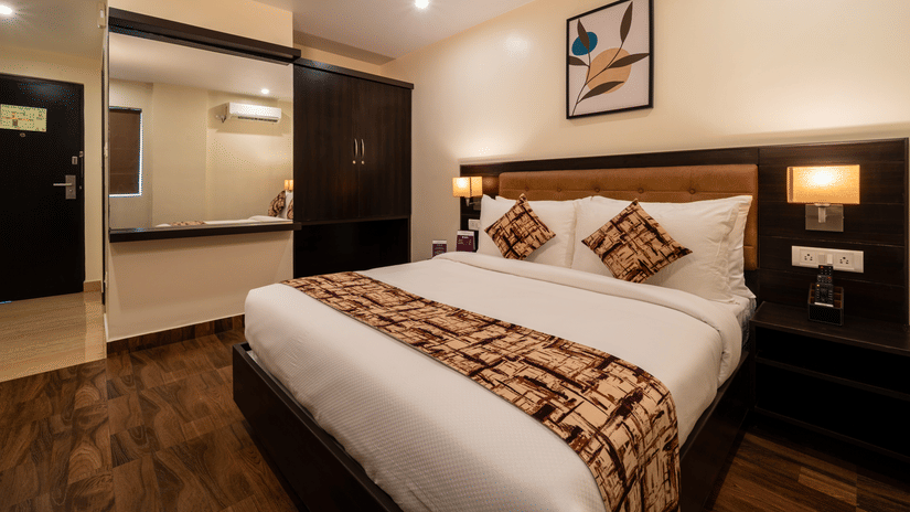 A bedroom interior at VITS Select, Bengaluru, featuring a plush bed, wooden flooring, and a bathroom entrance with mirror.