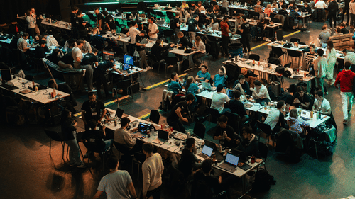 An expo where people are walking around inside a large indoor space in front of participants seated at long tables in various rows