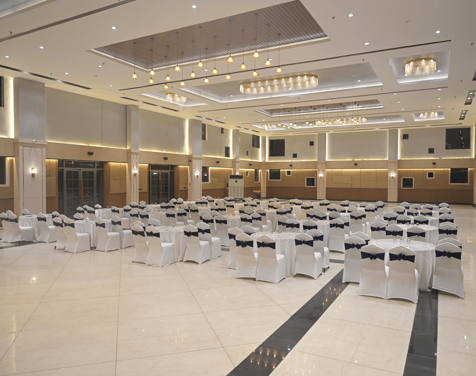 A panoramic view of the banquet hall arranged for a gathering, featuring bright chandeliers and neatly placed round tables at Hotel Sonar Bangla Mayapur.