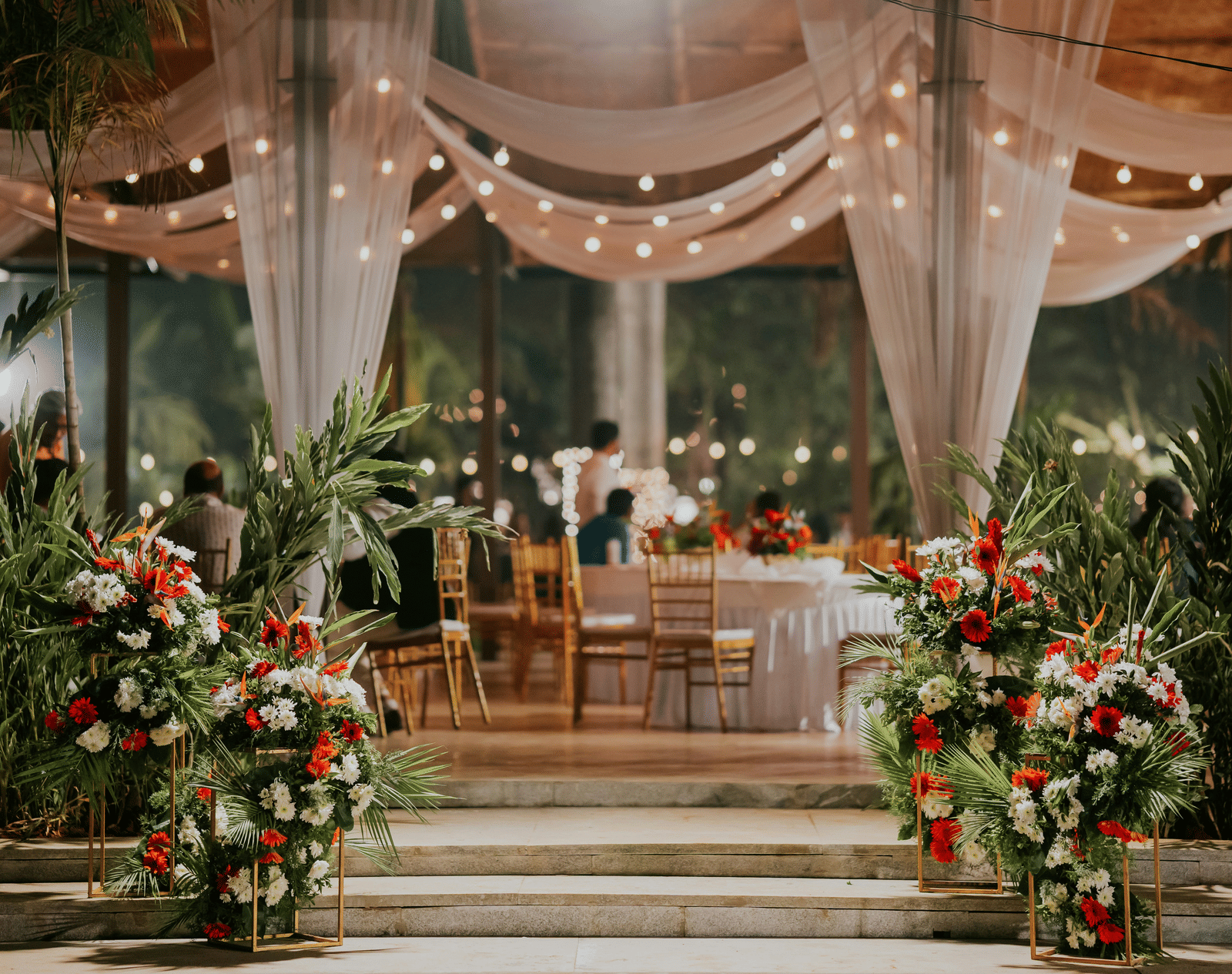 A beautifully decorated outdoor event space with white fabric draping, fairy lights, and large floral arrangements.