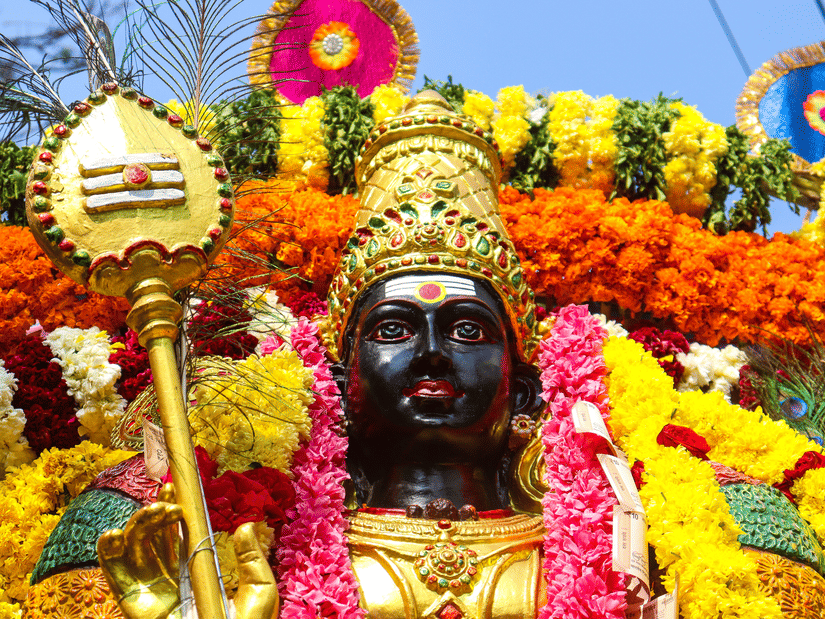 Close-up of a Lord Murugan idol decorated with bright yellow and orange marigold garlands during a festival celebration.