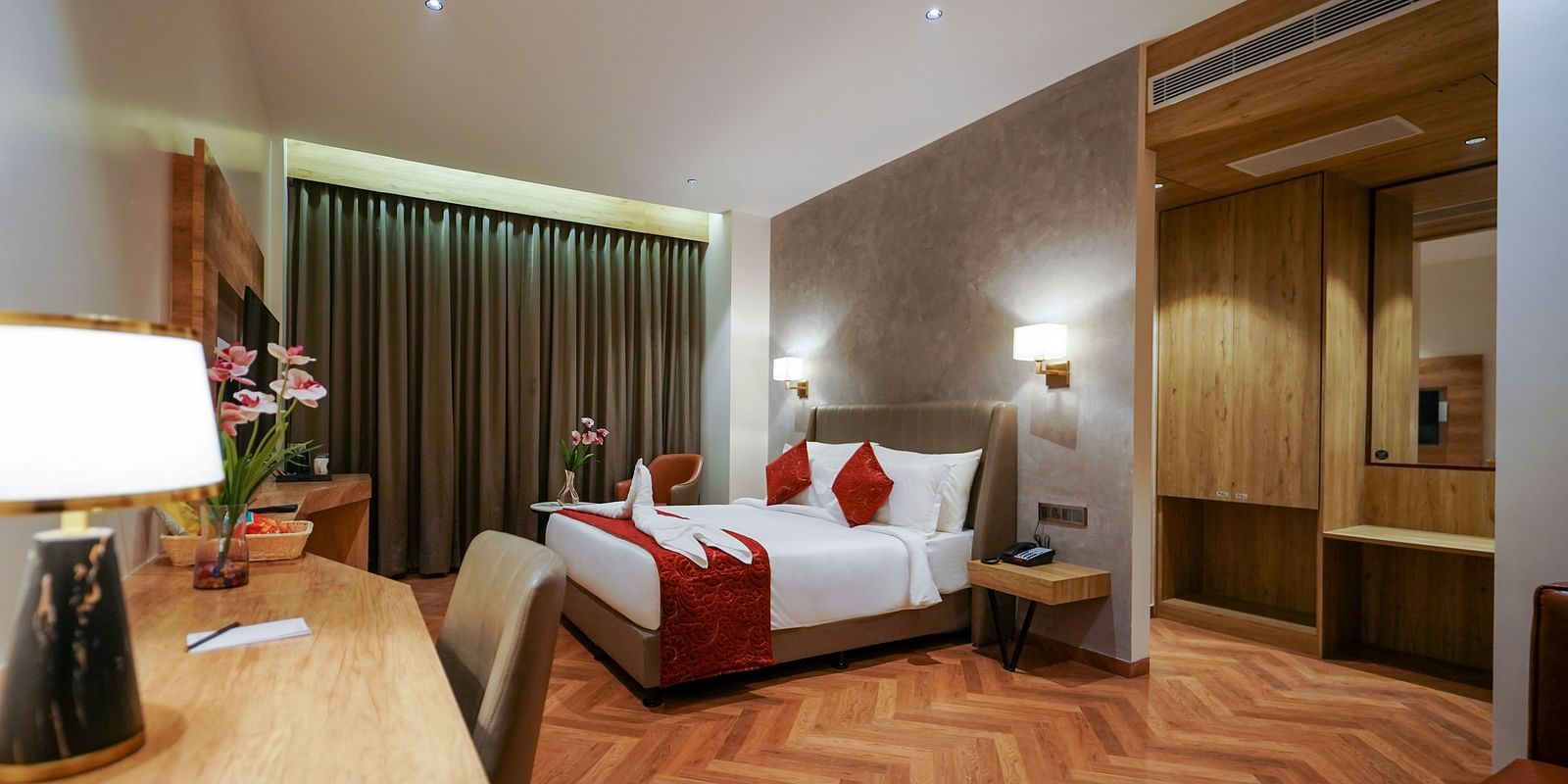 A well-lit hotel room includes a bed with pillows, a wooden desk with a lamp, a chair, curtains, and patterned flooring - Hotel Park Inn, Nellore.