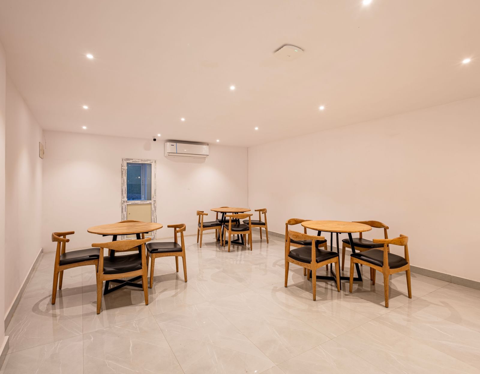 A brightly lit, minimalist dining or common area with several wooden tables and chairs arranged on a light-colored floor - Beestay Hotels Financial District, Hyderabad 