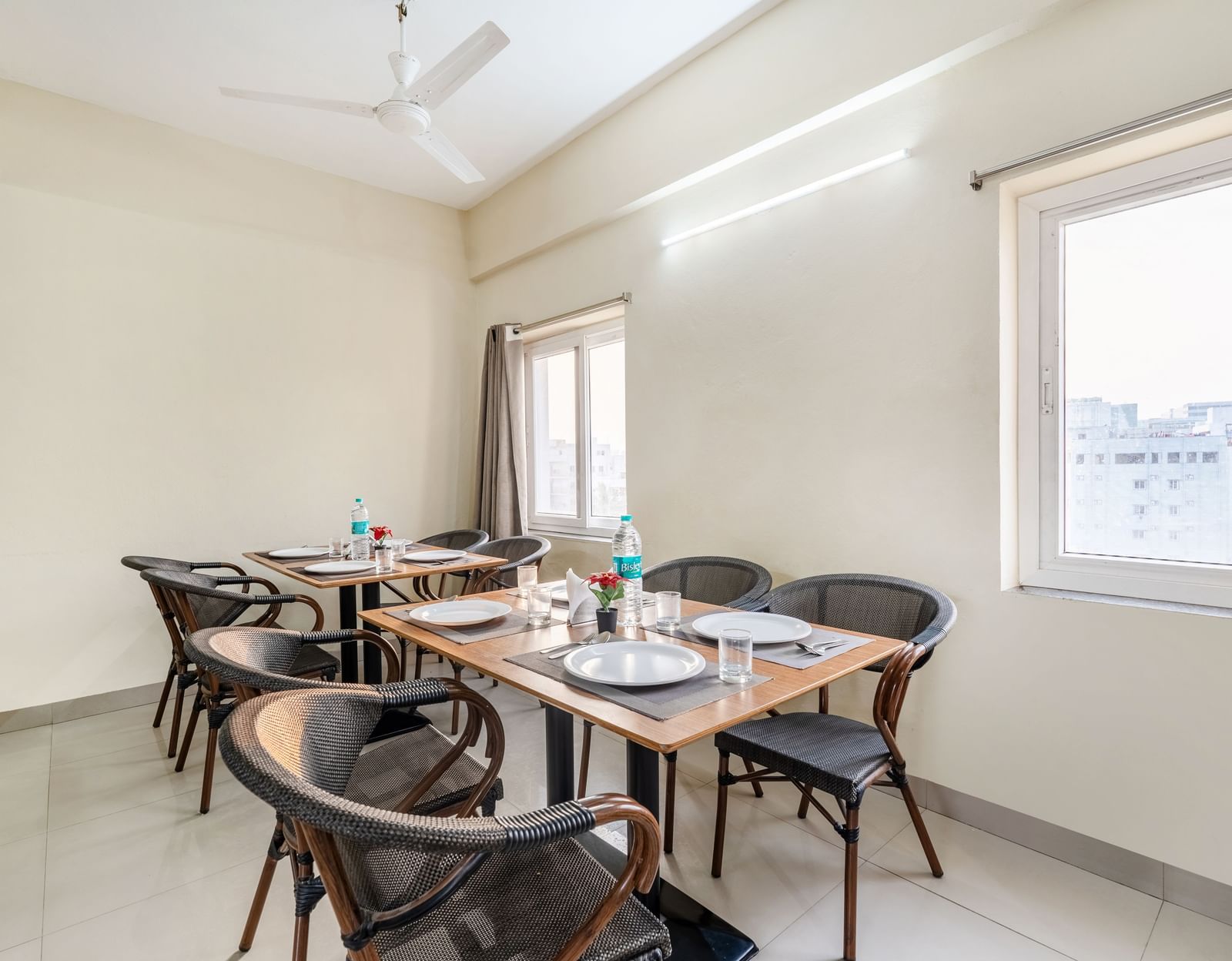 A close-up shot of a table in the dining area of Destino by Monday Hotels, Gachibowli, set with plates, glasses, and a bottle of water next to a window with grey curtains.