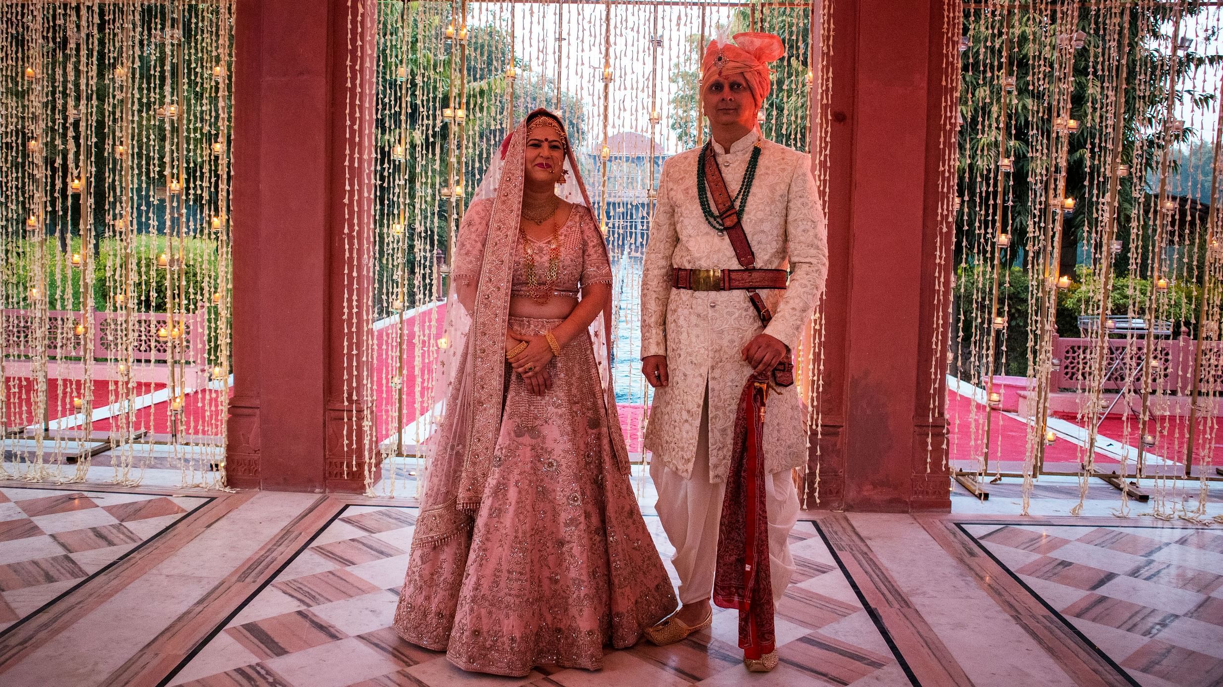 A bride and groom stand together in traditional outfits, framed by a curtain of hanging flowers- Umaid Palace - Best Luxury Heritage Resort Near Jaipur. 