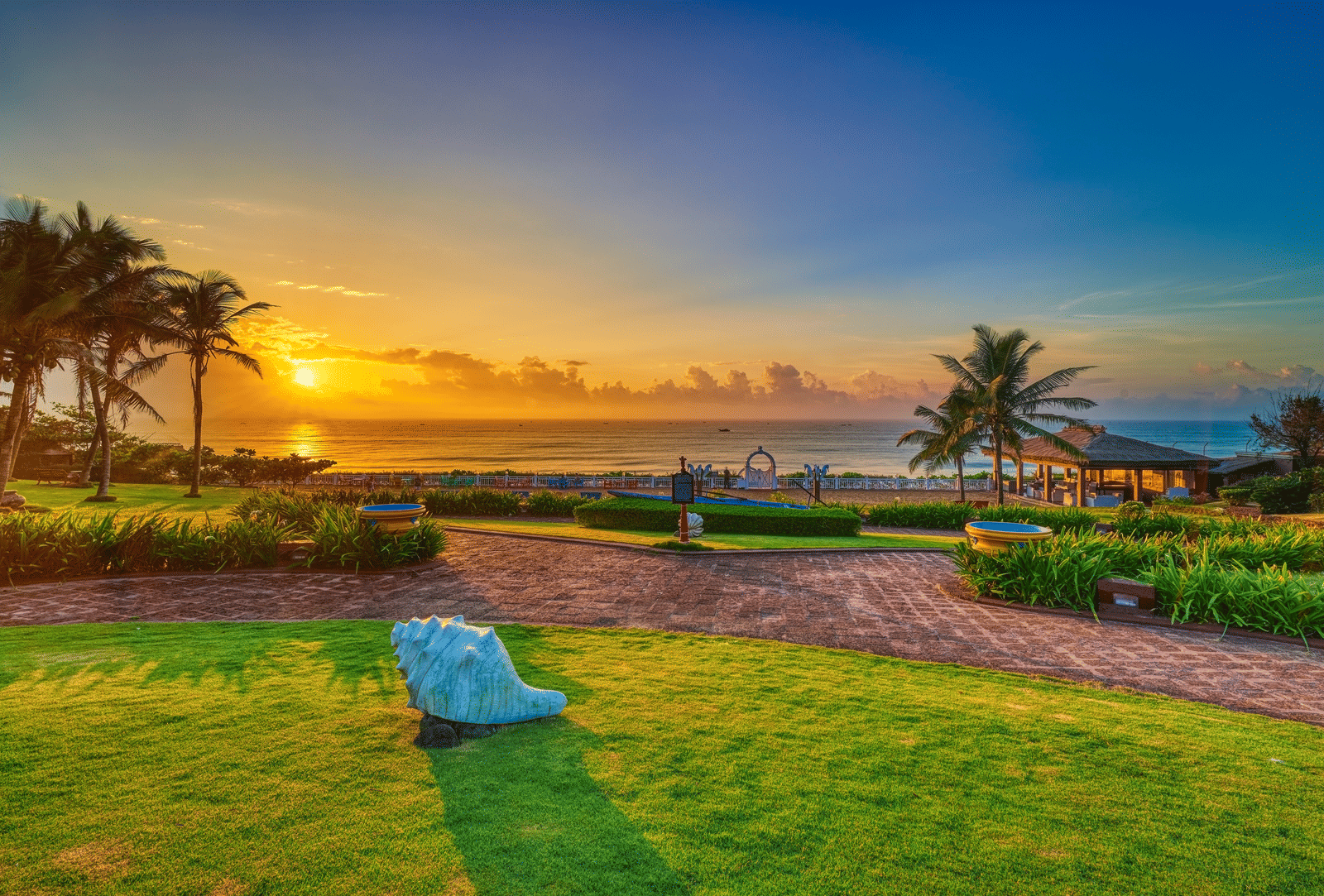 Seafront lawn at sunrise with a large conch-shell sculpture, palm trees, garden pathways and a gazebo at MAYFAIR Palm Beach Resort, Gopalpur-on-Sea.