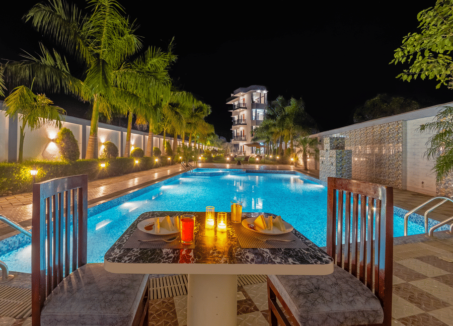 A romantic dinner table set for two beside a glowing blue swimming pool at night - Nature Trails Rishikesh